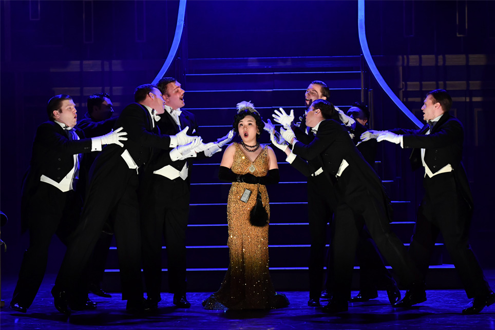 A women wearing a golden 1920s dress, singing on stage, with a group of men wearing smart historical suits and wearing gloves, surrounding her, with a dark blue staircase in the back
